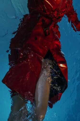 Mannequin en manteau rouge vif, immergé partiellement dans l’eau, mettant en valeur une esthétique audacieuse et avant-gardiste sous un éclairage aquatique sophistiqué.