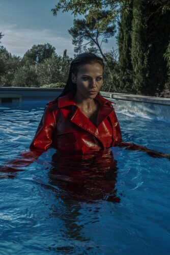 Mannequin portant un manteau rouge en vinyle, à la pointe de la mode, posant dans une piscine, ambiance moderne et élégante en extérieur.