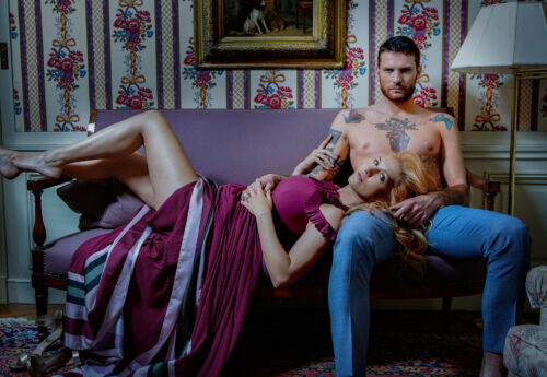 Deux mannequins posent élégamment sur un canapé ; la femme porte une robe longue bordeaux, l’homme torse nu affiche des tatouages, créant un contraste moderne et sophistiqué.