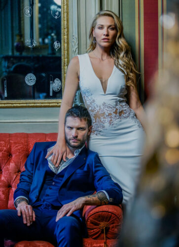 Deux mannequins posent élégamment, l’un en costume bleu satiné, l’autre en robe blanche ajustée à décolleté, dans un décor sophistiqué et raffiné.