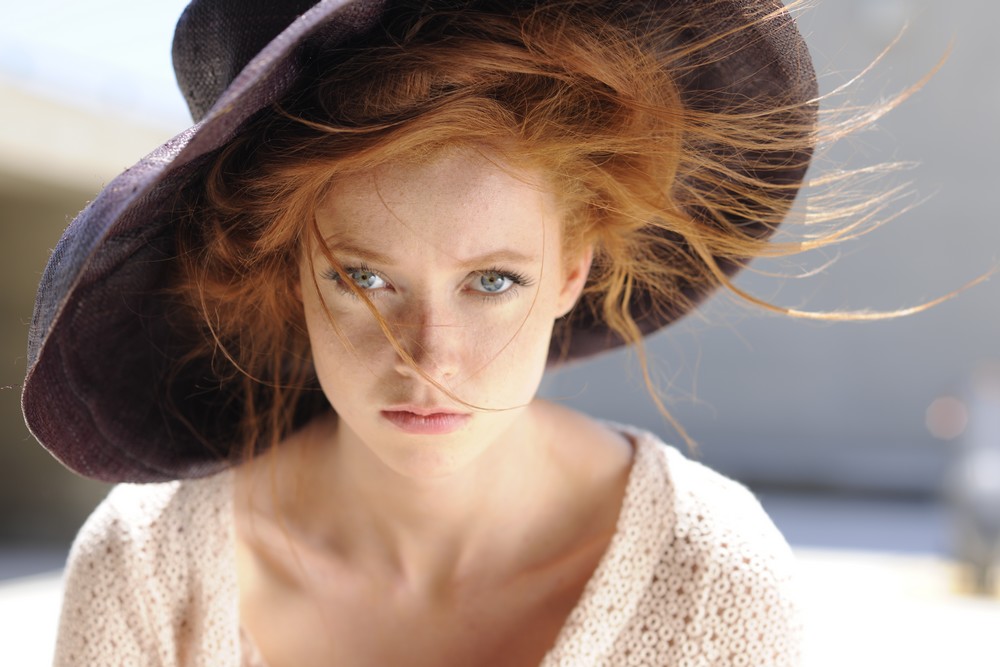 Mannequin aux cheveux roux portant un grand chapeau noir et un pull en maille crème, regard intense, style élégant et raffiné pour un éditorial de mode.