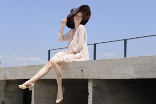 Mannequin portant une robe fluide beige et un grand chapeau noir, assise élégamment sur un muret, look estival sophistiqué, accessoires raffinés, pose gracieuse sous ciel bleu.
