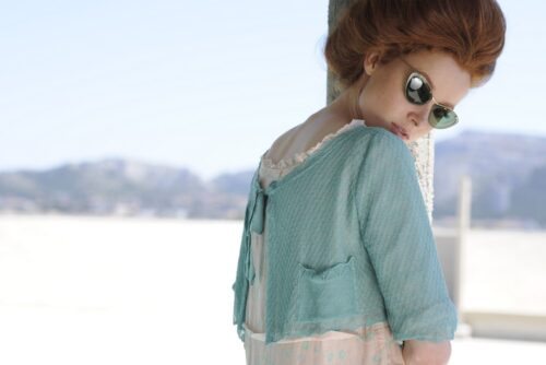 Mannequin portant une veste légère bleue pastel, lunettes de soleil rondes, coiffure volumineuse, style estival chic et décontracté en extérieur avec fond flou.