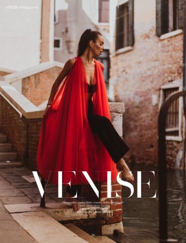 Mannequin portant une longue veste rouge fluide sur une combinaison noire, posé élégamment près d’un canal à Venise. Style sophistiqué, ambiance urbaine chic.