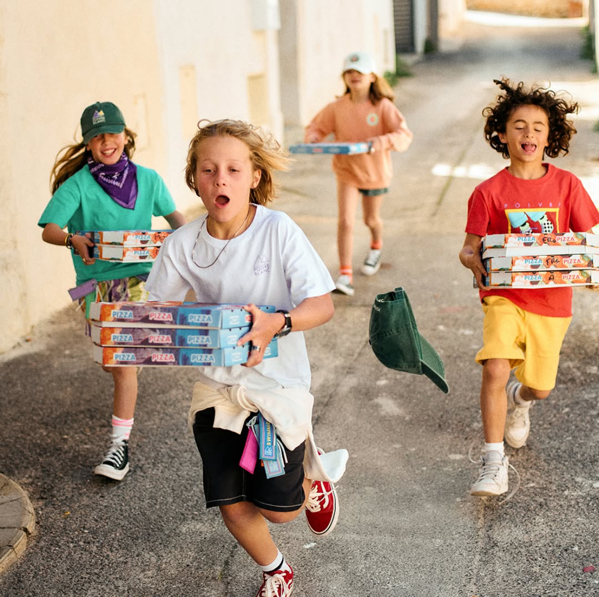 Quatre mannequins enfants portent des vêtements décontractés colorés, baskets et casquettes, courant avec des boîtes de pizza dans une ruelle ensoleillée, style urbain estival.