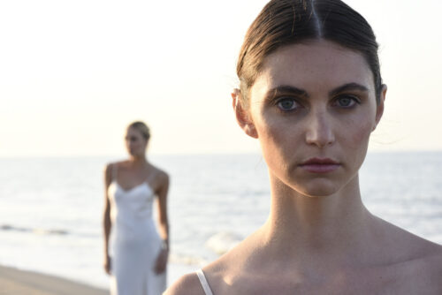 Deux mannequins en robes blanches élégantes posent sur une plage, mettant en valeur une esthétique minimaliste et sophistiquée, avec la mer en arrière-plan.