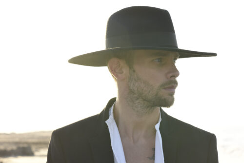 Mannequin masculin portant un grand chapeau noir et une veste chic, style élégant et décontracté, ambiance mode contemporaine sur fond extérieur lumineux.