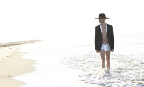 mannequin marche sur la plage, portant une veste noire, un chapeau large et un maillot blanc, créant un contraste élégant et audacieux avec le décor balnéaire.