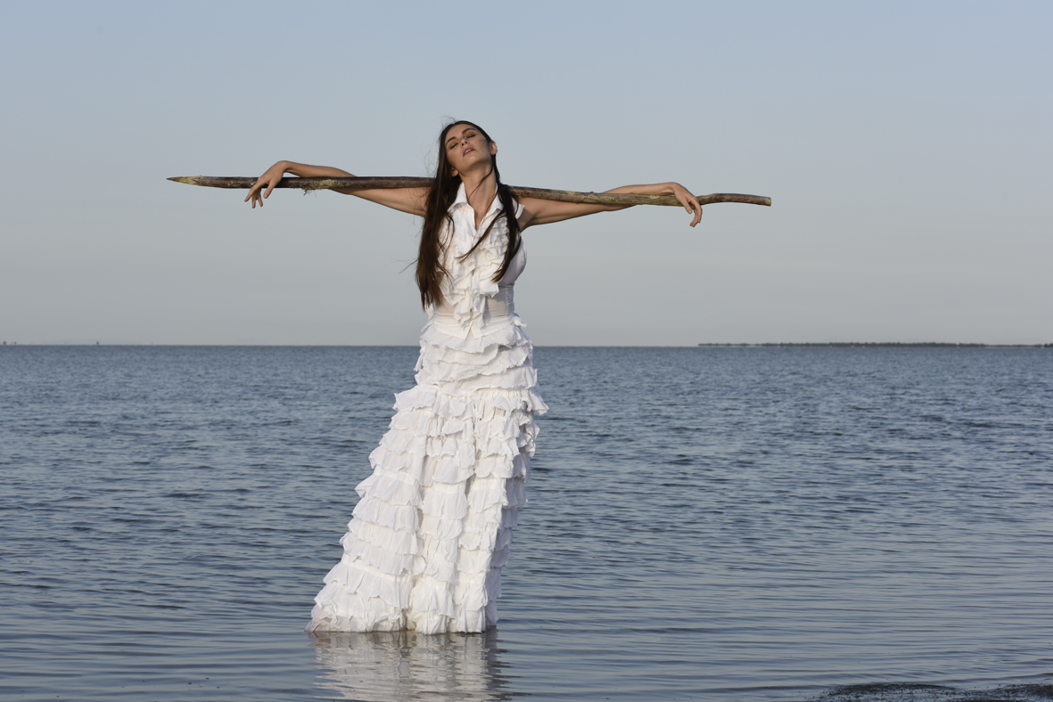 Mannequin portant une robe longue blanche à volants, posant de manière théâtrale dans l’eau, alliant élégance et créativité en extérieur.