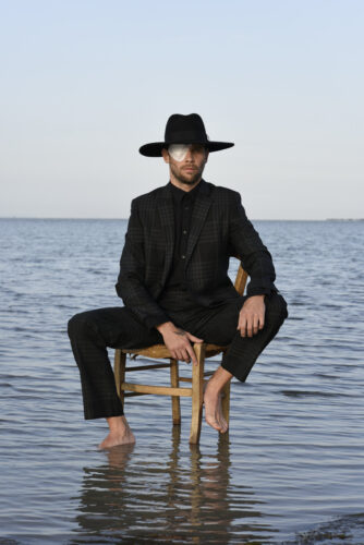 Mannequin habillé en costume noir élégant, assis pieds nus sur une chaise dans l’eau, portant un chapeau à larges bords et un accessoire argenté à l’œil.