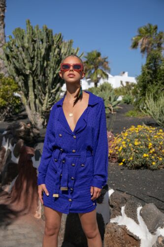 Femme portant une élégante tenue bleue Maison Monzerian, lunettes de soleil rouges, dans un jardin exotique avec cactus et fleurs, sous un ciel ensoleillé.