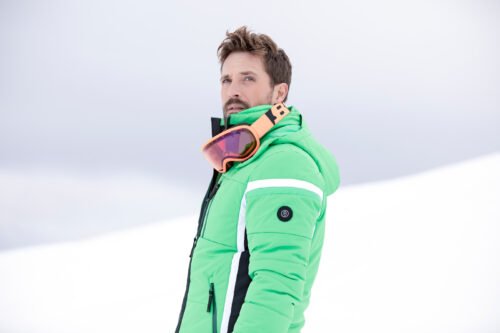Mannequin masculin portant une veste de ski verte tendance avec des détails noirs et blancs, accessoirisée de lunettes de ski orange, dans un décor enneigé, style sportswear moderne.