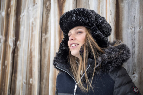 Mannequin portant une veste hivernale élégante et une chapka en fausse fourrure noire, posant devant un fond en bois pour une campagne mode hiver.
