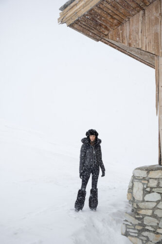 Mannequin en manteau d’hiver matelassé noir, pantalon assorti et bottes, pose près d’un chalet en bois sur fond de paysage enneigé, style hivernal sophistiqué.