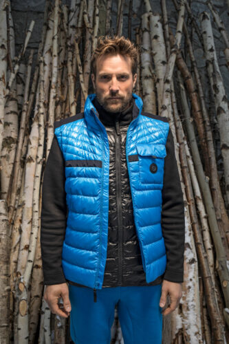 Mannequin homme portant une doudoune sans manches bleue ultra-moderne sur une veste matelassée noire, posé devant un décor de troncs naturels.