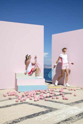 Deux mannequins posent dans un décor pastel, portant des tenues de sport blanches et roses, accessoires élégants, ambiance estivale et contemporaine, balles roses éparpillées au sol.