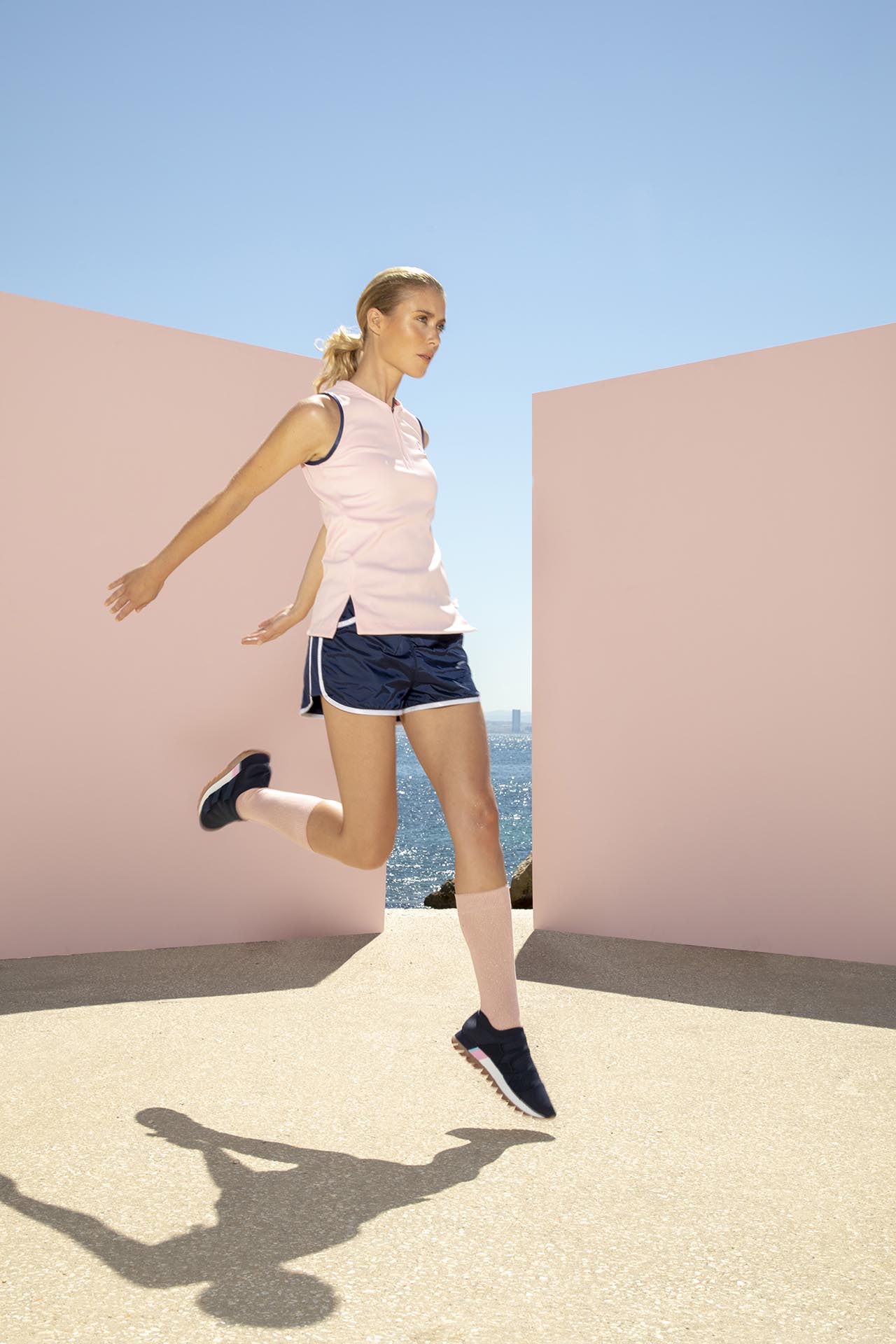 Mannequin en tenue de sport chic : polo rose pâle, short bleu marine, baskets noires, chaussettes montantes, ambiance minimaliste et estivale, décor pastel épuré.