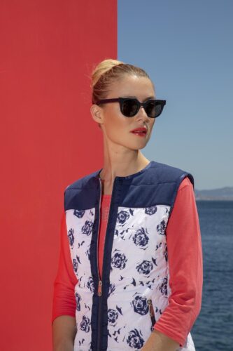 Mannequin portant des lunettes de soleil noires, veste imprimée bleu et blanc à épaules bleues, sur fond extérieur coloré, coiffure soignée et maquillage sophistiqué.