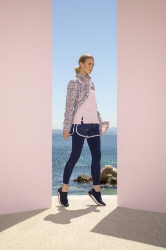 Mannequin en tenue sportive chic, veste imprimée, t-shirt rose pâle, short bleu et baskets, marchant en extérieur avec vue sur la mer, style décontracté et moderne.