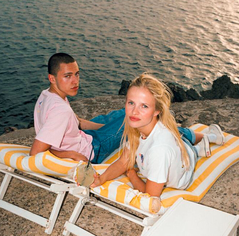 Deux mannequins posent avec décontraction sur des transats rayés, vêtus de t-shirts décontractés, créant un style estival moderne en bord de mer.