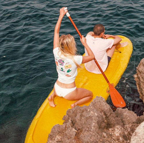 Mannequins portant des vêtements estivaux décontractés, t-shirt imprimé et maillot de bain blanc, posant sur un kayak jaune en bord de mer, capturant l’esprit estival tendance.