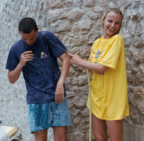 Deux mannequins arborent un style estival : t-shirt ample pour elle en jaune vif, t-shirt bleu et short pour lui, ambiance décontractée et ludique près d’un mur en pierre.