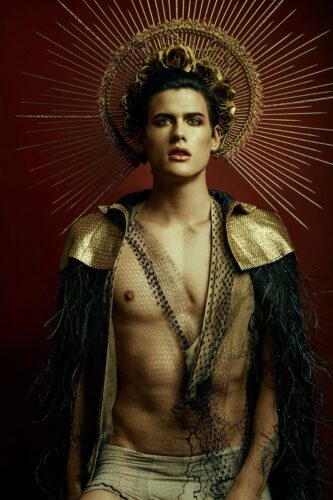 Homme debout, torse nu, auréole dorée, cape à plumes noires, arrière-plan rouge, style sophistiqué sous la direction artistique de Mimmo Carabetta.