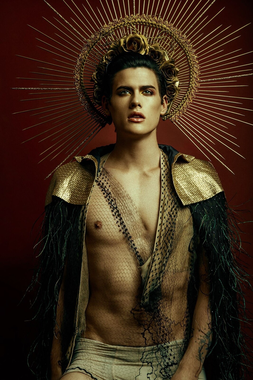 Homme debout, torse nu, auréole dorée, cape à plumes noires, arrière-plan rouge, style sophistiqué sous la direction artistique de Mimmo Carabetta.