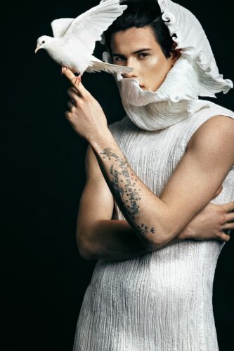 Portrait artistique d’une personne élégante en blanc, avec un tatouage, tenant une colombe, mettant en valeur la direction artistique de Mimmo Carabetta, sur fond sombre.