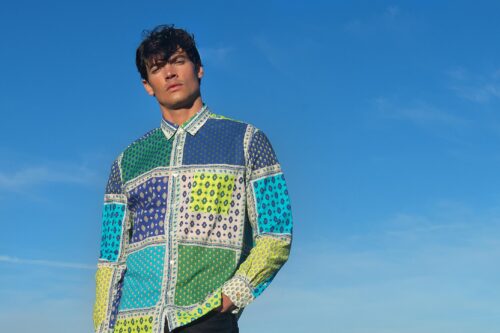 Mannequin portant une chemise patchwork aux motifs géométriques colorés, posant devant un ciel bleu, style contemporain et tendance décontractée.
