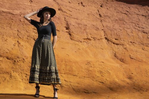 Mannequin portant un chapeau noir, une jupe longue imprimée, un haut noir et des sandales à talons, posé devant une falaise orangée, style bohème chic et élégant.