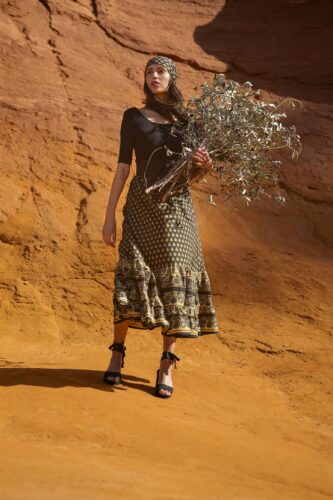Mannequin portant une robe midi fluide à motifs, sandales noires à talons, tenant un bouquet sec, posé sur un fond naturel ocre, ambiance élégante et bohème.