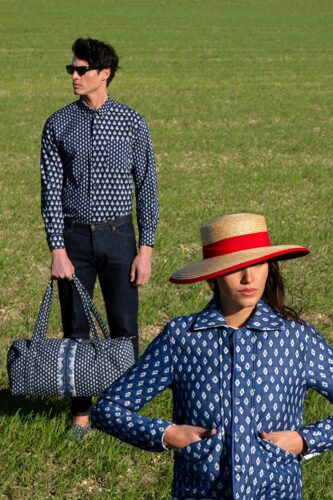 Deux mannequins portent des ensembles coordonnés bleu à motifs, dans un décor champêtre ; la femme porte un chapeau de paille élégant, l’homme tient un sac assorti.