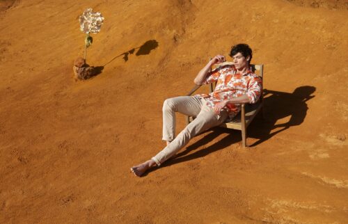 Mannequin habillé d’une chemise imprimée orange tropical et d’un pantalon beige, assis de façon décontractée sur une chaise sur fond de sable, ambiance mode estivale sophistiquée.