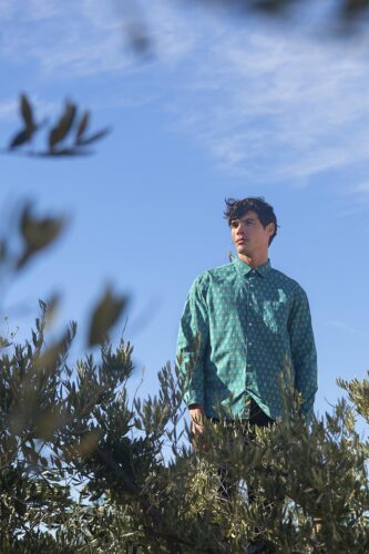 Mannequin homme portant une chemise verte à motifs, posé dans un décor naturel sous un ciel bleu, style décontracté et tendance, idéal pour une collection printemps-été.