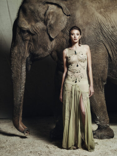 Mannequin en robe longue élégante beige et fendue, posant devant un éléphant, créant un contraste saisissant entre mode sophistiquée et nature sauvage.
