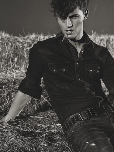 Mannequin masculin en chemise en jean foncé, ceinture à clous, posant de façon sophistiquée devant des bottes de foin, ambiance western moderne et élégante.