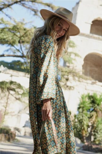 Mannequin portant une robe longue à motifs géométriques, manches longues et chapeau, dans un décor extérieur estival sophistiqué.