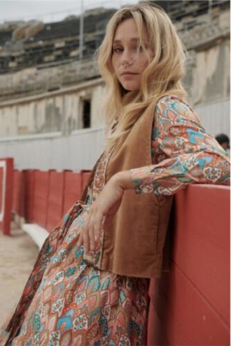 Mannequin portant une robe longue à motifs floraux et un gilet camel, posant dans une arène, style bohème chic, ambiance décontractée et élégante.