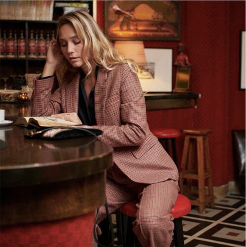 Mannequin en tailleur pantalon rose poudré, assise élégamment dans un bar raffiné, lisant un livre, ambiance sophistiquée aux tons rouges et mobilier vintage.