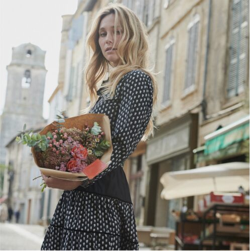 Mannequin habillée d’une robe à pois noire et blanche, tenant un bouquet de fleurs, pose dans une rue ancienne, ambiance élégante et citadine.