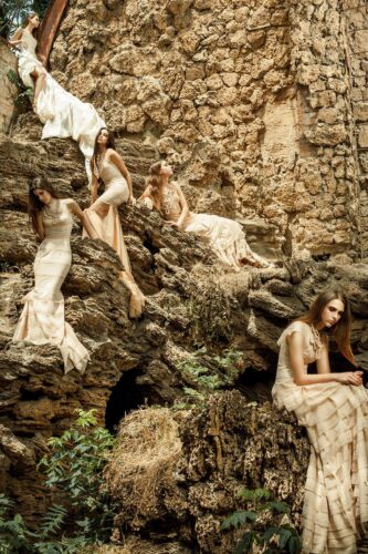 Des mannequins posent sur des rochers, portant des robes longues et élégantes de style bohème, dans un décor naturel et rustique, mettant en valeur la délicatesse des tissus.