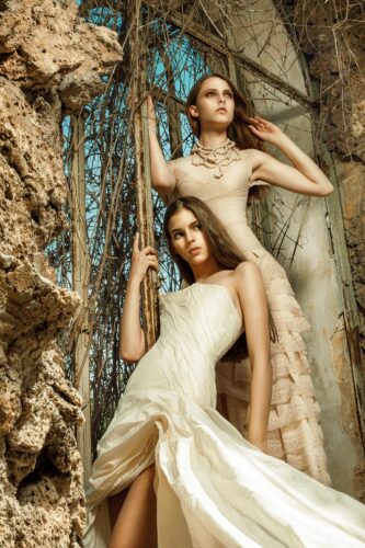 Deux mannequins posent dans des robes de haute couture ivoire et beige, au style élégant et sophistiqué, au milieu d’un décor naturel texturé avec des pierres et des branches.