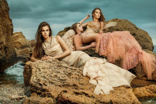 Trois mannequins posent sur des rochers, vêtues de longues robes élégantes aux tons neutres et pastel, mettant en valeur une esthétique sophistiquée et naturelle sous un ciel dramatique.
