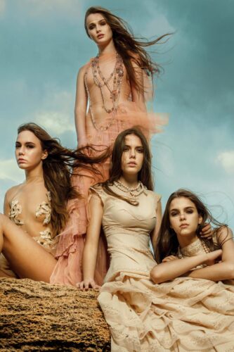 Quatre mannequins posent en extérieur, vêtues de robes élégantes aux teintes naturelles et aux textures fluides, incarnant un style sophistiqué et bohème sous un ciel nuageux.