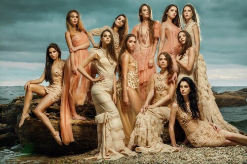 Groupe de mannequins posant en tenues sophistiquées couleur chair et beige, robes longues et étoffes transparentes, ambiance élégante et fashion sur un décor naturel.