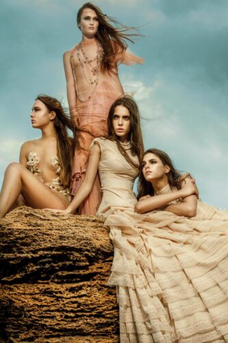 Trois mannequins posent sur des rochers, vêtues de robes fluides et élégantes aux tons neutres, inspirant une ambiance éthérée et sophistiquée sous un ciel bleu.