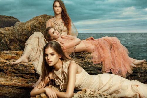 Trois mannequins posent sur des rochers en robes longues et fluides, style bohème chic, ambiance naturelle et élégante près de la mer.